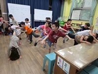 低年級學生跟著幕做出單腳站立，手腳均張開的動作，有個小女生單腳站，雙手扶地板，另一腳抬高